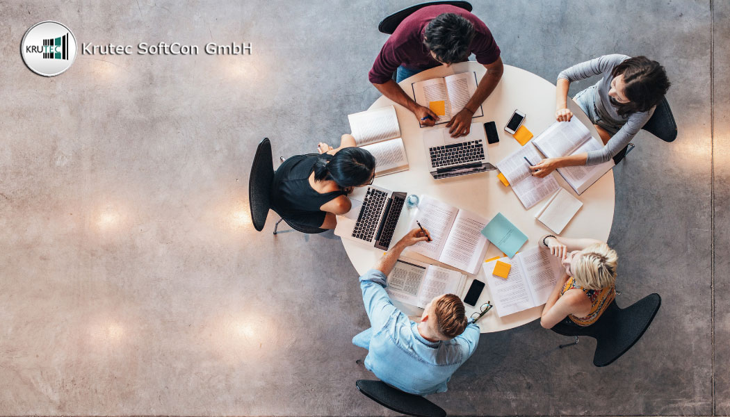 Projektteam sitzt an einem runden Tisch und arbeitet mit Laptops, Smartphones, Büchern und Post-its