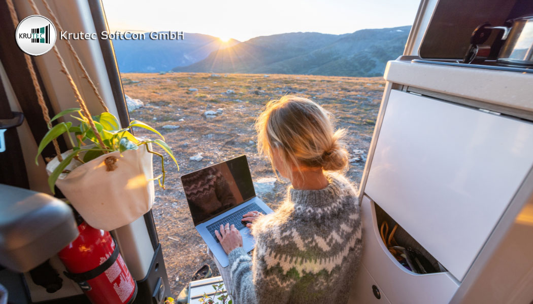 Mitarbeiterin in Remote Work arbeitet in der Tür ihres Campers am Laptop mit Blick auf eine Berglandschaft in ihrer Umgebung